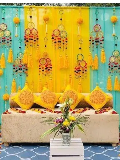 A beautiful outdoor Mehandi seating area with a backdrop of turquoise and yellow drapes. The sofa is decorated with printed cushions, and the setup includes unique hanging tassels.
