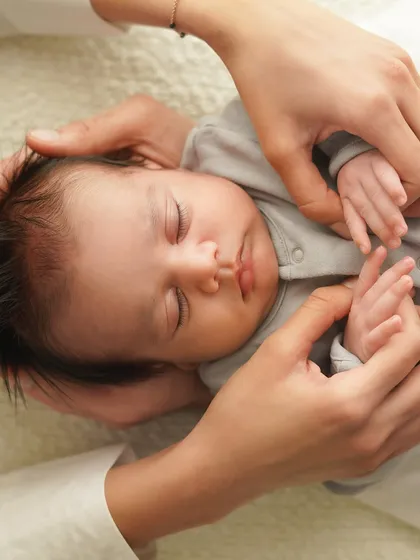 Parents' hands frame their sleeping baby's face, a gesture of love and protection. It's a quiet, intimate moment frozen in time.
