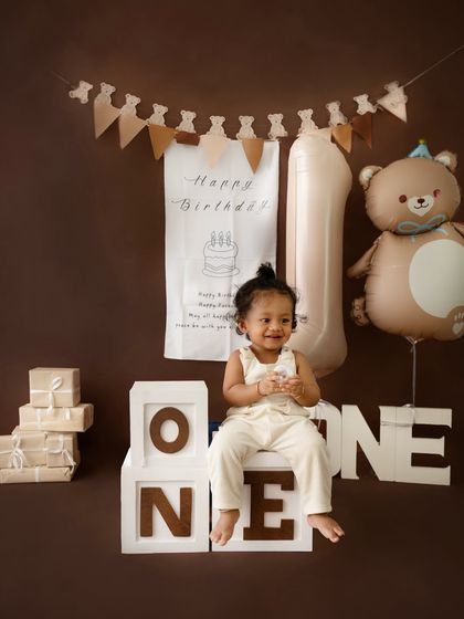 A joyful first birthday photo with a warm, brown-toned teddy bear theme. The setup includes a large "1" balloon, gift boxes, and a happy baby boy.