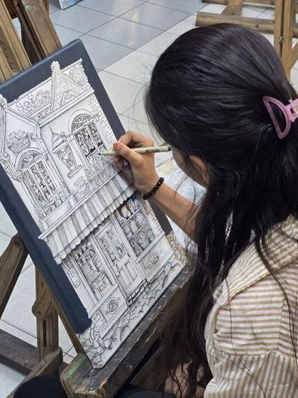 An over-the-shoulder view of a student working on her architectural drawing with a micron pen, carefully adding details to the windows and facade.