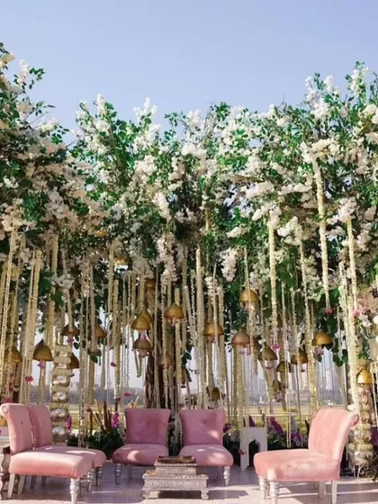 A mandap designed to feel like a forest clearing, with tall floral structures and a canopy of hanging jasmine and temple bells. The soft pink chairs add a touch of modern elegance.