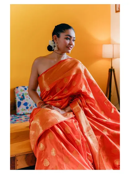 An intimate, lifestyle shot that feels both candid and elegant. The model is seated in a warmly lit room, showcasing an orange silk saree in a comfortable, natural setting.