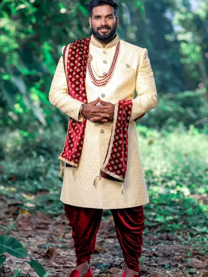 A happy client looking absolutely majestic for his outdoor wedding ceremony. He is wearing a custom-tailored cream sherwani with a maroon velvet stole, paired with traditional dhoti pants and red shoes.