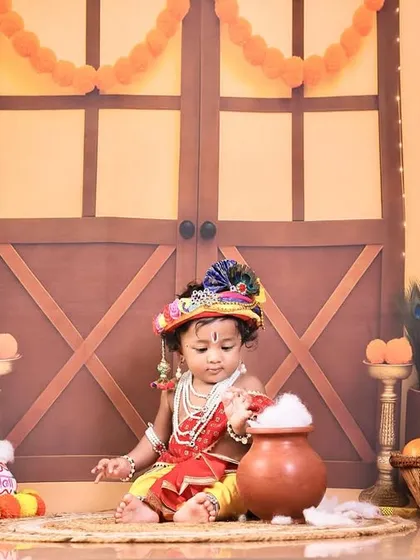 A candid shot of our little Krishna curiously playing with the "butter" in a pot, set against a rustic, village-themed backdrop.