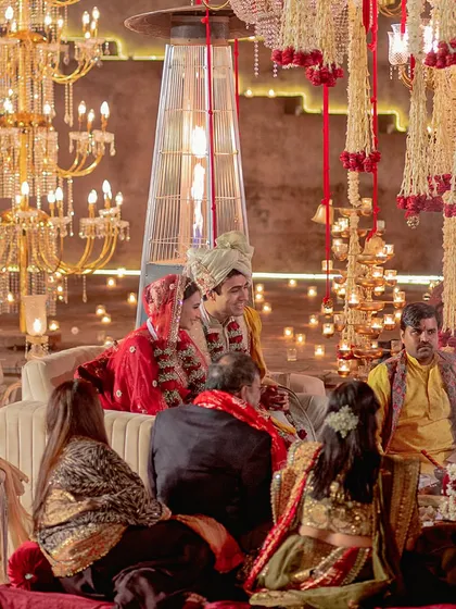 A couple partakes in their wedding rituals, seated under a canopy of hanging floral strings and surrounded by the golden light of grand chandeliers and candles.