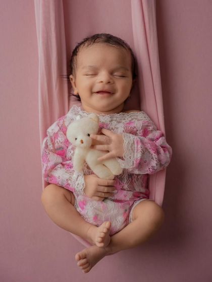 A smiling newborn in a floral pink outfit cuddles a tiny white teddy bear while resting in a soft pink sling.