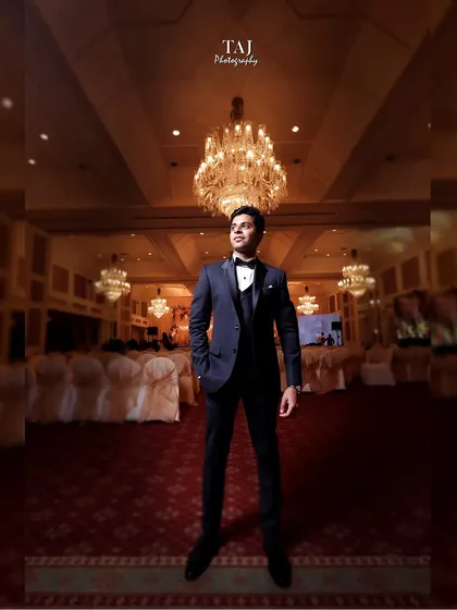 A dapper groom standing tall in a grand ballroom. The low-angle shot emphasizes his sharp tuxedo and the magnificent chandeliers above.