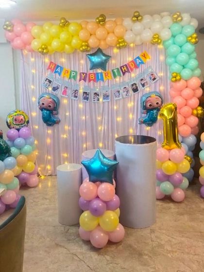 A Cocomelon themed birthday decoration for a home party. The setup includes a curtain backdrop with fairy lights and colorful balloon pillars.