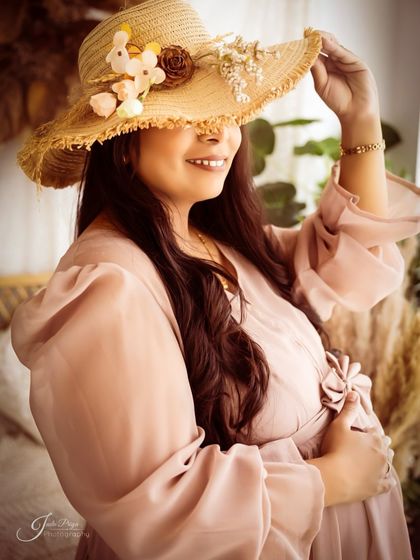 A playful and chic portrait using a straw hat as a prop. The mom-to-be peeks out from under the hat with a smile, adding a touch of rustic charm to her look.