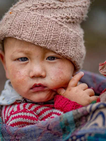 A close-up portrait of a baby with rosy cheeks, bundled up against the cold in Himachal. The child's curious gaze is captivating.