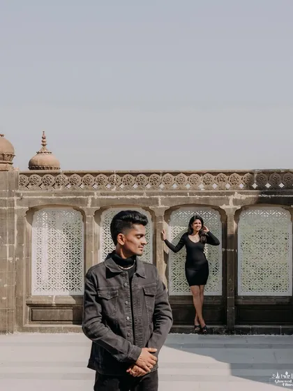 A creative composition focusing on depth and perspective. The groom stands in the foreground while the bride poses against the palace wall, creating a stylish, editorial-style pre-wedding photograph.