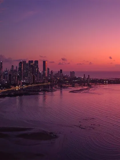 A panoramic view of the Mumbai coastline and the Bandra-Worli Sea Link, bathed in the purple and pink light of a dramatic sunset.