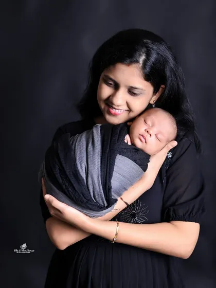 A mother's gentle smile radiates love as she cradles her sleeping newborn. This classic portrait uses a dark background to draw all attention to the peaceful and loving bond between mother and child.