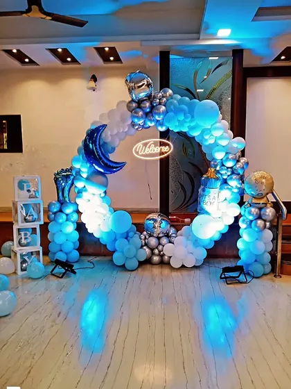 A circular balloon arch in blue and silver for a celestial-themed welcome party. The design includes a metallic blue moon, a 'Welcome' neon sign, and other space-themed balloon accents.