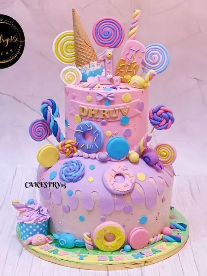 A fantastical two-tier Candyland-themed cake. This cake is a sugar paradise, covered in fondant lollipops, donuts, macarons, and ice cream cones for a first birthday.