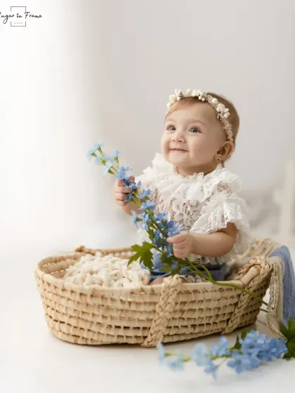 A baby girl in a wicker basket, holding a sprig of blue flowers that perfectly match her bonnet and romper.