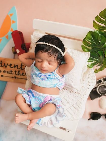 A newborn dreams of the beach. This creative setup features a baby girl in a tiny swimsuit, relaxing on a mini beach chair with a surfboard and camera props.
