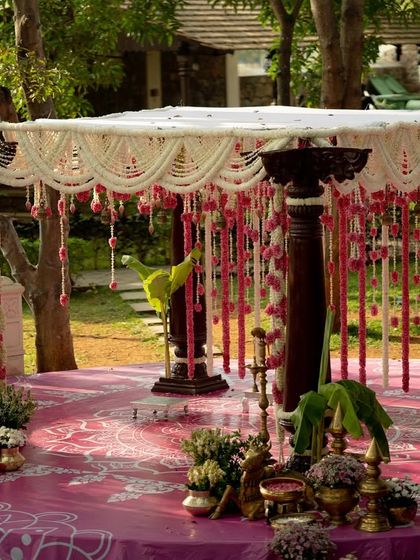 A closer look at the outdoor wedding mandap. The structure is built with traditional dark wood pillars, and the roof is a delicate net of jasmine flowers, from which hang beautiful strings of pink carnations and lotus buds.