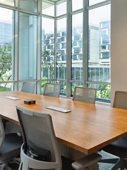 A minimalist conference room flooded with natural light from floor-to-ceiling windows. The simple wood table and ergonomic chairs create a clean, calm, and productive environment.