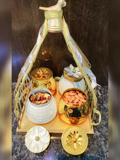 A golden metal basket holding four ornate jars, two in white ceramic and two in gold metal, filled with dry fruits and treats.