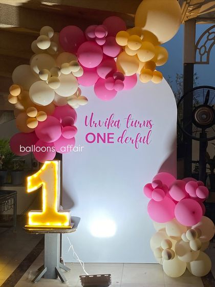 A sweet and simple 'Onederful' first birthday setup. The decor features a pink and peach balloon garland on a white arched backdrop with a light-up number '1'.