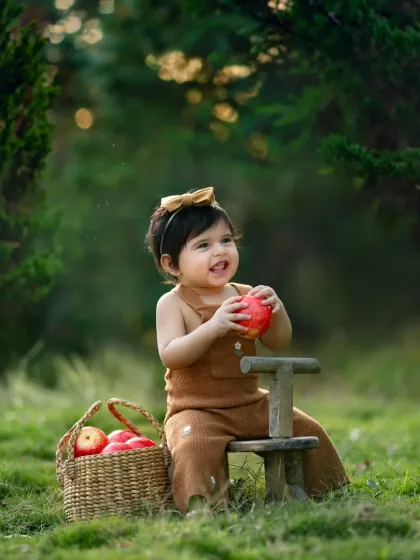 A toddler sits on a small wooden bicycle, holding an apple from a nearby basket. A charming and rustic-themed outdoor shoot.