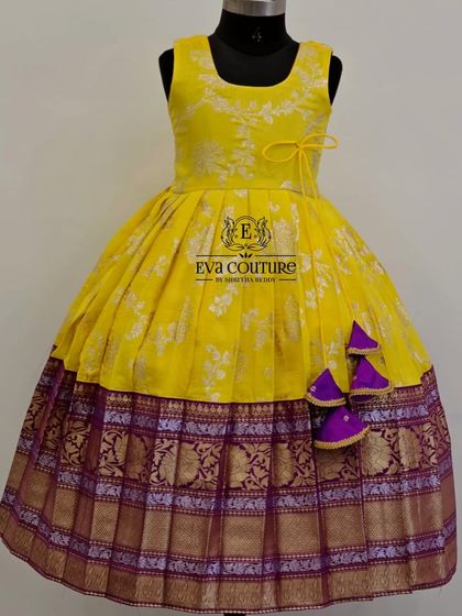 A sleeveless yellow brocade frock with a contrasting purple Kanchi Pattu border. The cute tassels add a pop of color.