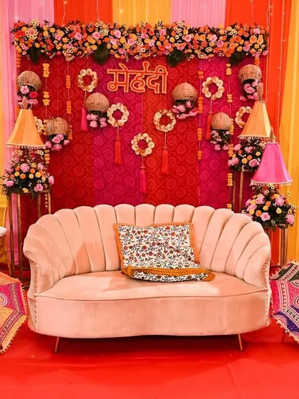 A classic mehendi seating setup with a vibrant pink and yellow backdrop, traditional Rajasthani umbrellas, and a plush velvet sofa.