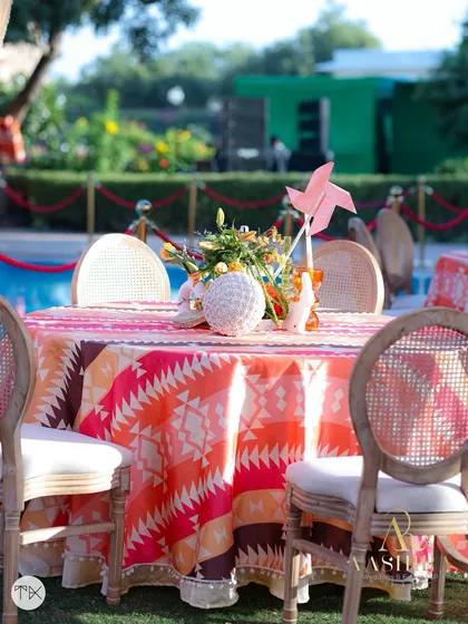 A poolside table with a vibrant patterned tablecloth and a centerpiece featuring a pink pinwheel.