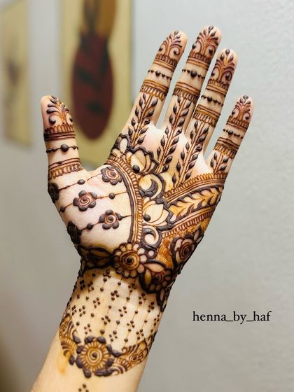 An intricate bridal henna design with a mix of floral and paisley elements. The palm features a large, detailed motif with hanging flowers, while the fingers are covered in leafy vines.