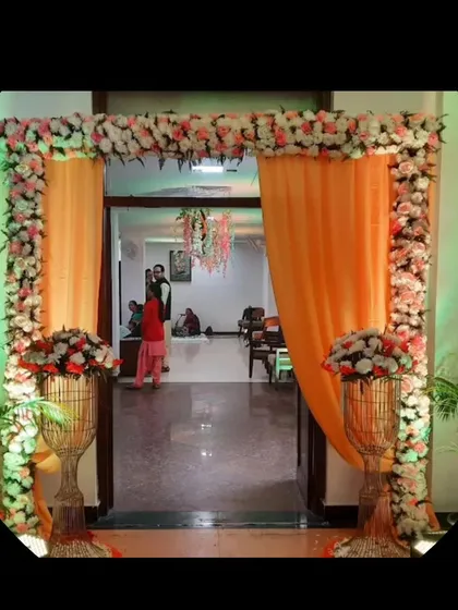 An indoor entrance decoration for a home function. The doorway is framed with an arch of pink and white flowers and orange drapes, creating a beautiful transition between spaces.