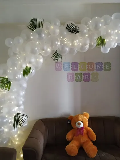 A simple and elegant 'Welcome Baby' decoration with a white balloon arch intertwined with fairy lights and green leaves.