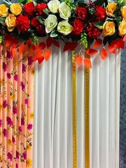 A simple backdrop decoration that combines a pleated white curtain with a colorful floral-printed fabric panel. The top is adorned with a mix of artificial flowers and leaves, showing how different materials can be used for a custom look.