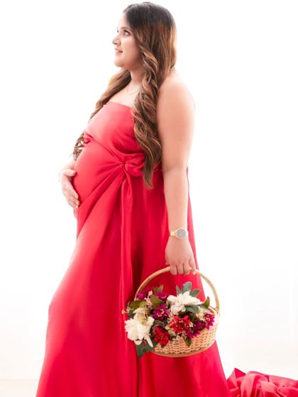A side profile that emphasizes the elegant drape of the red gown and the beautiful curve of the baby bump. The clean background keeps the focus entirely on the mother's form.