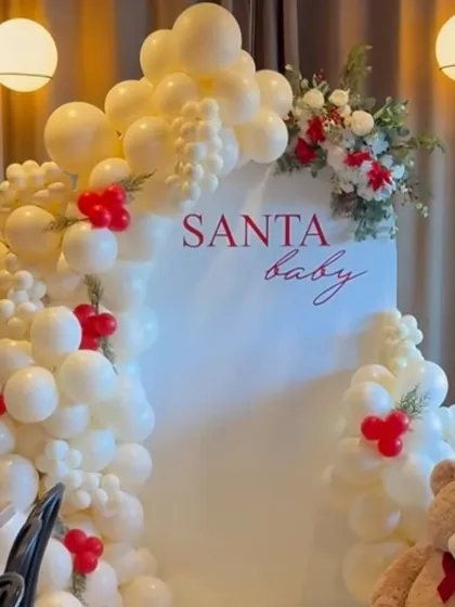 An elegant and soft Christmas theme, perfect for a 'Santa Baby' baby shower or party. This setup uses a white arch panel and a cloud of white balloons with small red accents and floral touches.