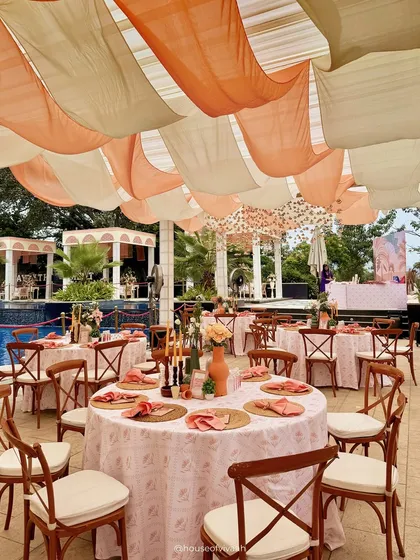 An outdoor dining area by the pool, with tables dressed in pastel linens and surrounded by wooden cross-back chairs, all under a billowing canopy of peach and white fabric.