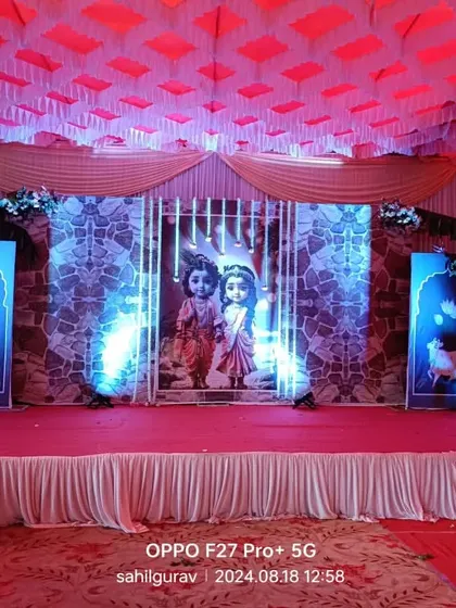 A stage decoration for a Krishna-themed event. The backdrop features an image of Radha and Krishna, with Pichwai-style cow cutouts and dramatic blue and red lighting.