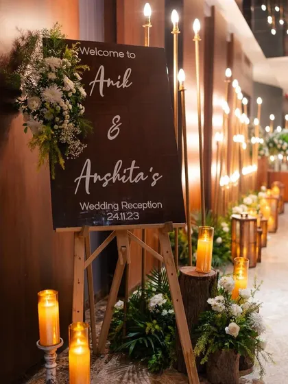 A rustic wooden welcome sign on an easel, flanked by candles on log stumps, creating a warm and inviting entrance for a reception.