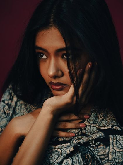 A more contemplative shot from the same series. The model's pose, with hands framing her face, feels thoughtful and a little melancholic. The rich burgundy background adds to the moody, operatic feel of the editorial.