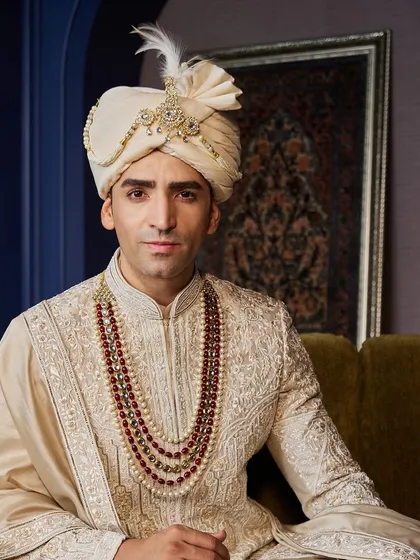A portrait of regal elegance. This Angarkha-style sherwani in a soft cream color is adorned with a stunning multi-layered ruby and pearl necklace, creating a look of pure opulence.