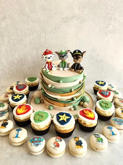 A full dessert spread for a Paw Patrol fan. The cake is surrounded by themed macarons and cupcakes, each featuring the logo of a different pup.