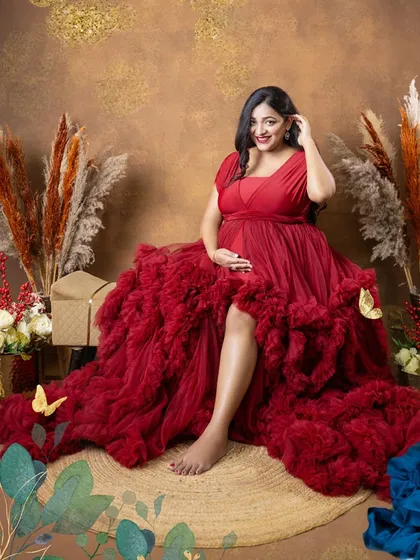 A relaxed and confident solo portrait. The rustic, bohemian-style set provides a warm and earthy backdrop for the striking red ruffled gown.