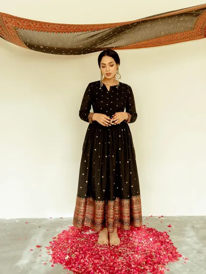 A full-length portrait for Safaa World. The model stands on a carpet of red petals, creating a strong color contrast with the black and gold anarkali.