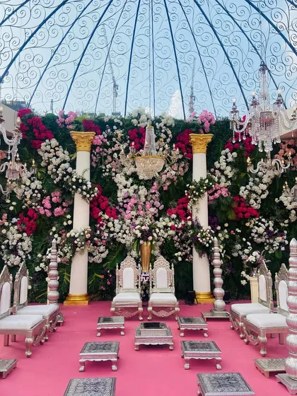 A direct view of the floral mandap stage, framed by a glass dome. The wall of flowers in shades of pink, white, and red creates a stunning backdrop, complemented by Roman-style pillars and elegant silver seating for a royal feel.