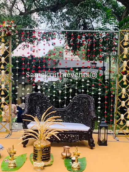 A full view of a traditional outdoor engagement stage. The setup includes a dark carved bench, brass lamps, and decorative panels with hanging jasmine and bells.