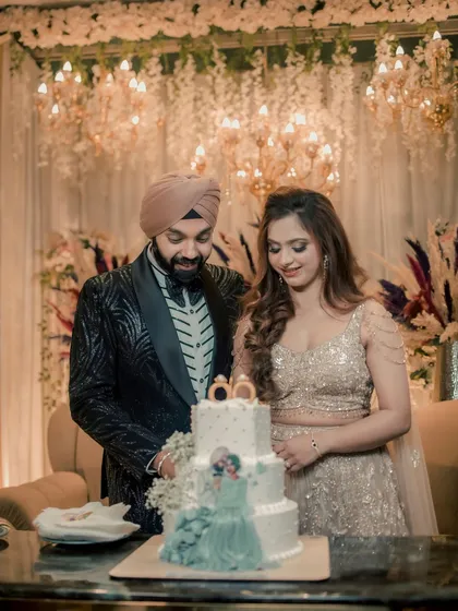 The couple, Simran and Sanjit, share a moment of joy as they cut their tiered engagement cake, surrounded by a beautiful floral and chandelier-lit backdrop.