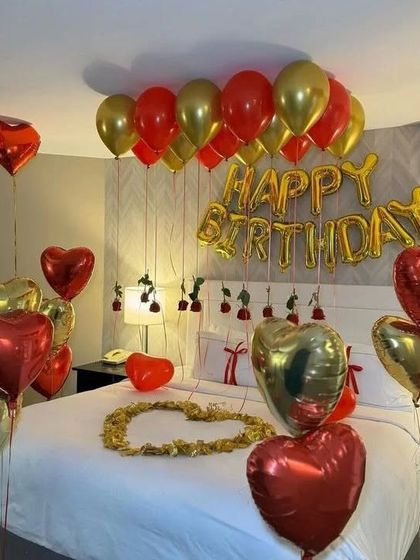 A romantic birthday setup in red and gold, with heart-shaped foil balloons, "Happy Birthday" letters, and a heart of petals on the bed.