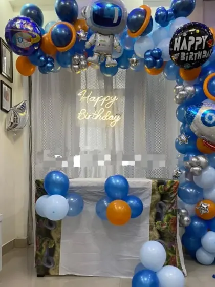 A space-themed birthday decoration perfect for a home setting. The balloon arch features an astronaut, a rocket, and planets, with a "Happy Birthday" neon sign in the center.