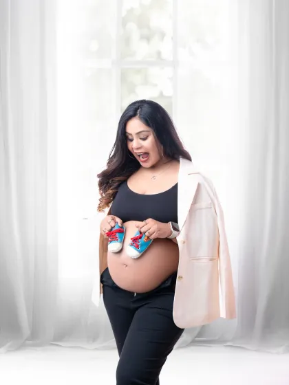 A modern and chic announcement. The mother-to-be, in a stylish blazer and crop top, excitedly holds up a tiny pair of sneakers.