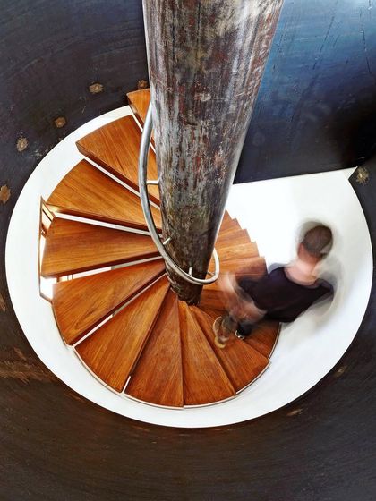 A person descends a spiral staircase, their motion captured as a blur. The design of the stair, with its solid central column and radiating wooden treads, creates a powerful sense of movement and descent.